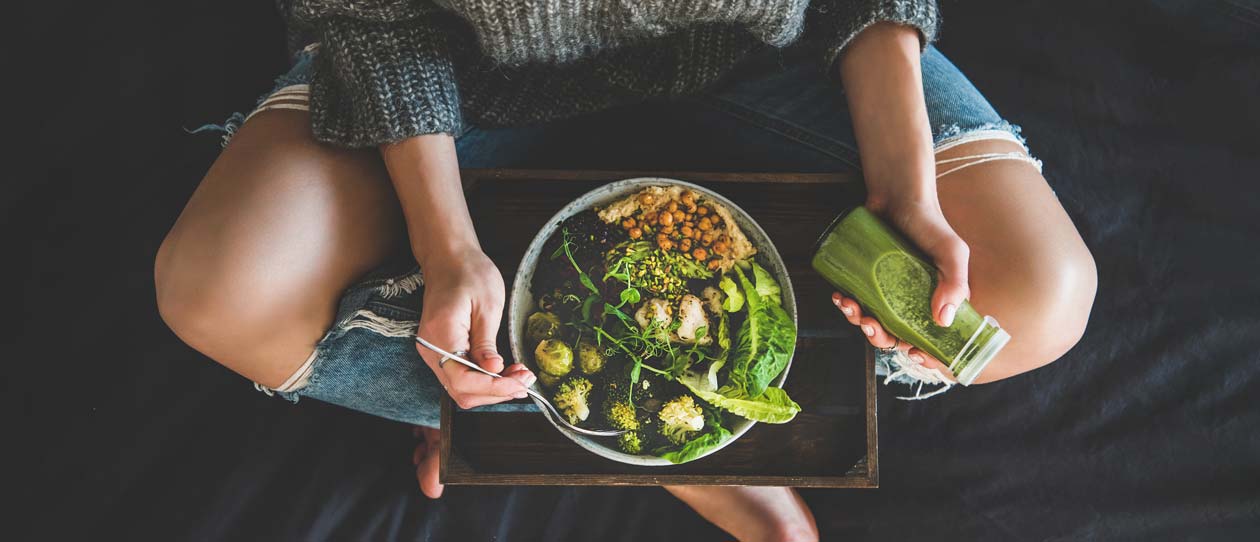 Healthy dinner, lunch in bed. Woman sitting in bed, eating vegan superbowl or Buddha bowl with hummus, vegetable, fresh salad, beans, couscous and avocado and drinking green smoothie, top view