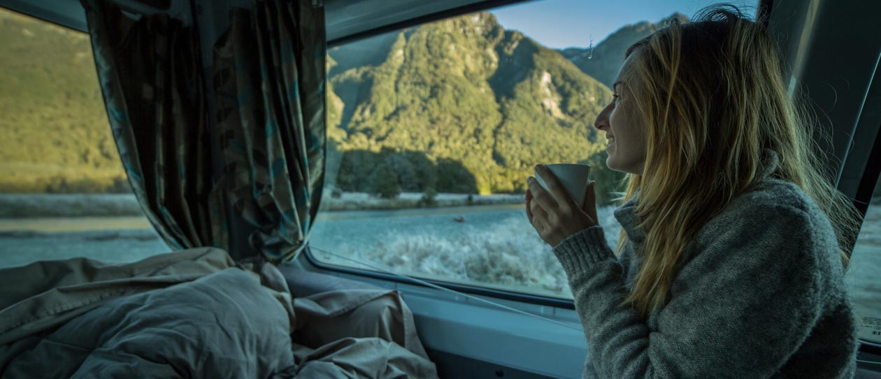 Young woman in a camper van drinks a tea and looks through window at a beautiful mountain landscape on an early winter morning.