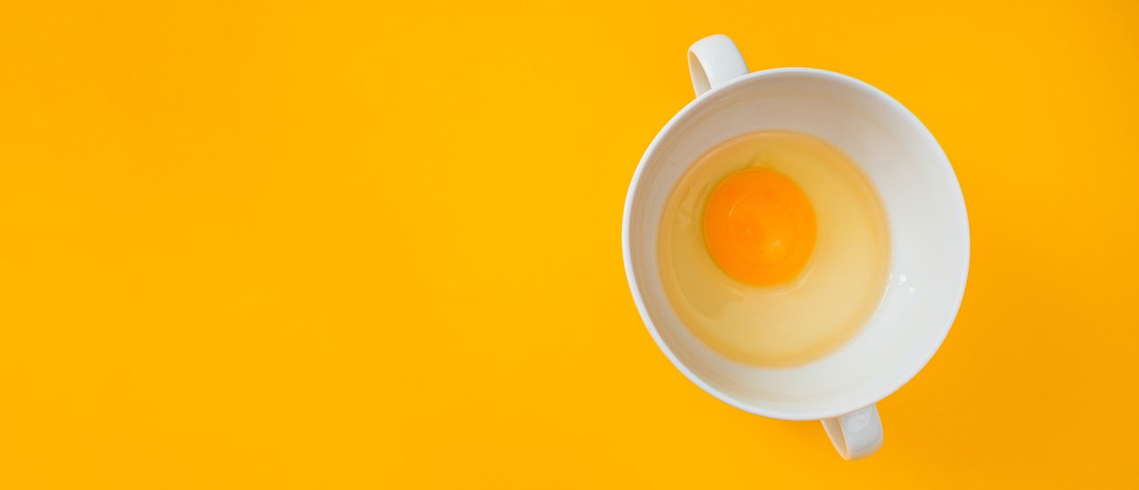 One raw egg in a white cup on a yellow background