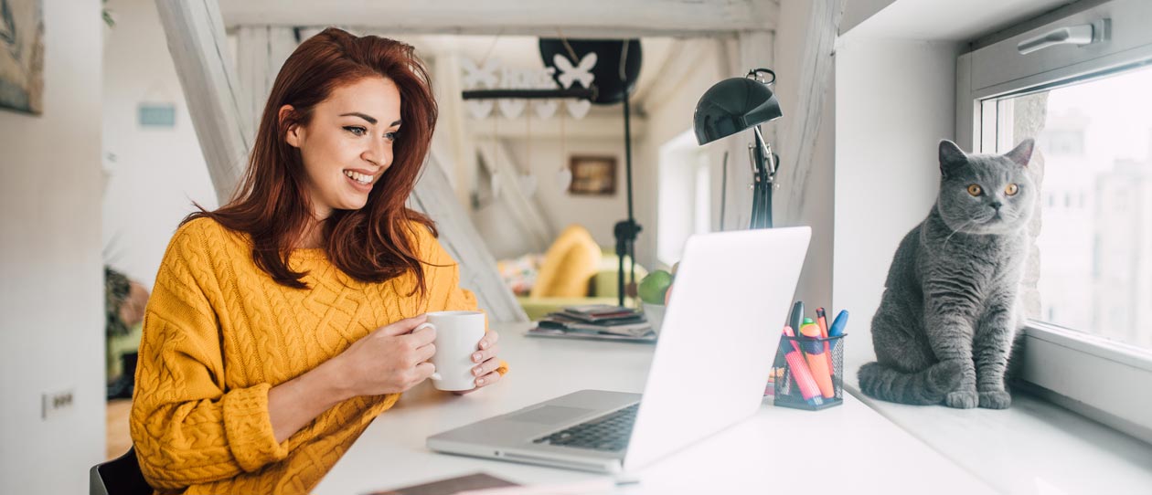 Smiling young redhead holding a cup of coffee while working on her laptop in her home office with a cat sitting in a window